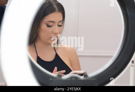 A woman is sitting in front of a lighted mirror, looking at her phone. The mirror is circular and the woman is wearing a black top Stock Photo