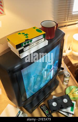 Stacked VHS tapes with old CRT television and assortment of vintage media equipment surrounding it. Includes coffee cup and blinds in the dimly lit scene Stock Photo