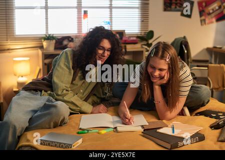 Students sitting on bed, engrossed in collaborative learning. Books, notebooks, and stationery scattered on bed, creating a productive and casual study atmosphere Stock Photo