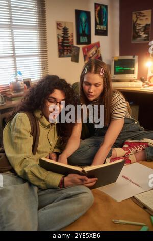 Engaged students sharing ideas while reading book together in cozy room with posters and vintage computer in background for focused study environment with attentive expressions Stock Photo