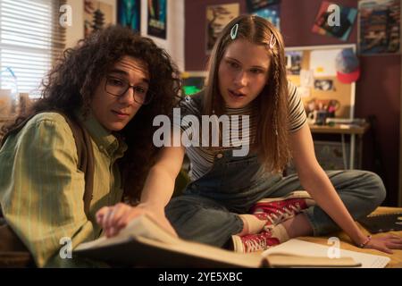 Two teenagers studying together in home office, focusing on book. Warm lighting illuminates cozy room with posters and decorations visible in background Stock Photo