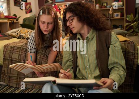 Two teenagers separately sitting on couch, examining notebooks and writing notes, engrossed in study. Background shows subtle room decor and soft lighting reflecting focused atmosphere Stock Photo