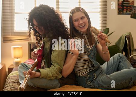 Young couple sitting on bed, smiling while playing guitar and enjoying music. Natural light from window gives cozy and warm retro ambiance to scene Stock Photo