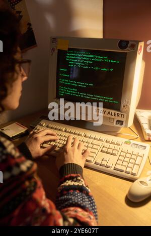 Individual working on programming project with a retro desktop computer displaying code on screen in dimly lit room using a desk lamp, keyboards and other tech devices visible Stock Photo