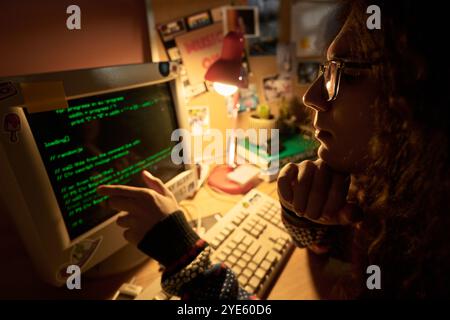 Young programmer engrossed in coding on an old-style computer in a cozy, warmly lit home office setting filled with various retro decorations and a desk lamp Stock Photo