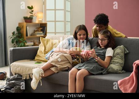Three children sitting on gray sofa, looking at tablet with concentration, surrounded by home decor and warm lighting. Cozy atmosphere in modern living room Stock Photo