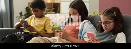 Group of three children sitting on sofa, each focused on their mobile device. Diverse group featuring two girls and one boy, all deeply engaged in their screens Stock Photo