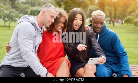 Cheerful multiethnic male and female friends taking selfie through smart phone while sitting in public park. Happy men and women and enjoying leisure time together outdoors. Stock Photo