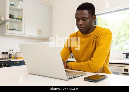 African american man in kitchen using laptop, wearing yellow sweater, smartphone on counter, at home Stock Photo