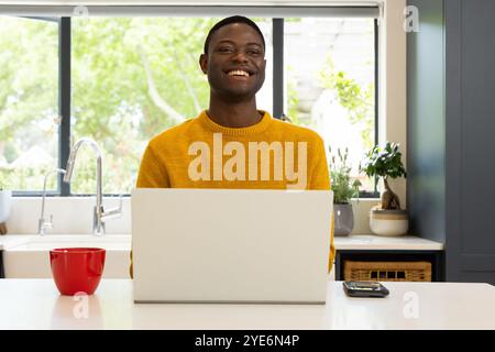 Smiling African american man in yellow sweater using laptop at home with red mug nearby Stock Photo