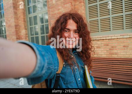 Happy non-binary student taking a selfie outdoors, smiling confidently in a casual setting with long curly hair, denim jacket, and backpack, capturing a personal moment outside an academic building. High quality photo Stock Photo