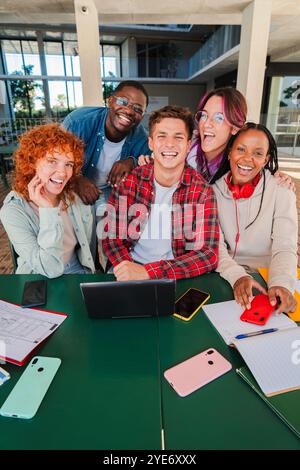 Vertical. Smiling multiracial group of young students looking at camera, sitting together in university library with a laptop, books, and notebooks. Diverse classmates collaborating at university. High quality photo Stock Photo