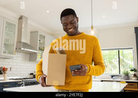 Smiling African american man in kitchen checking smartphone while opening delivery box, at home Stock Photo