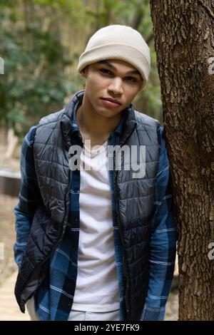 Young man in beanie leaning against tree in forest, looking relaxed Stock Photo