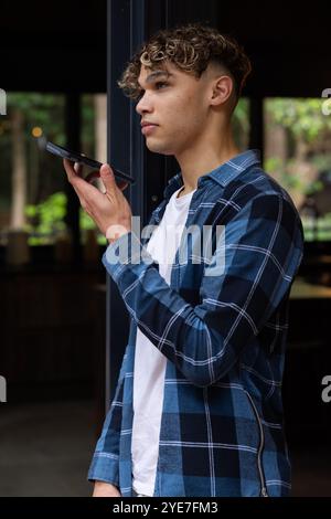Young man using smartphone voice assistant at home, wearing casual plaid shirt Stock Photo