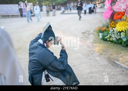 Man in a black hat and a blue jacket is taking a picture with his cell phone. He is sitting on the ground Stock Photo