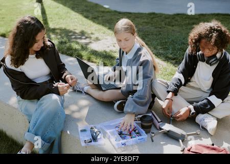 Biracial teen friends in casual clothes sitting on cement making drone together while using laptop to read instruction Stock Photo