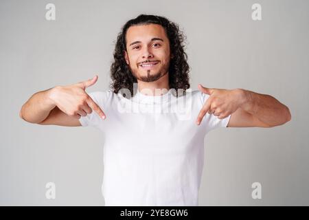 A young man with curly hair joyfully pointing down while wearing a plain white t-shirt against a neutral background Stock Photo