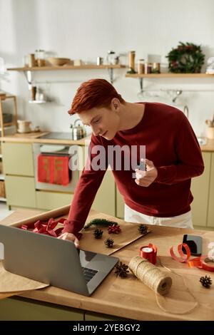 The young man is busy decorating for Christmas while using a laptop in his kitchen. Stock Photo
