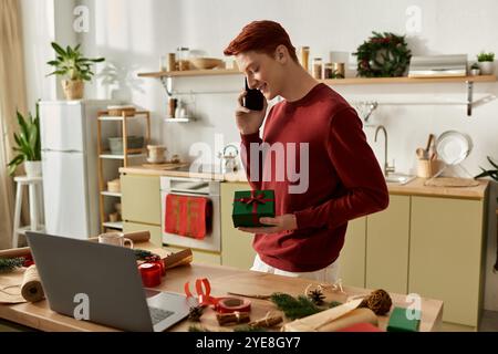 The young man holds a gift and smiles during a festive phone call in a warm, decorated kitchen. Stock Photo