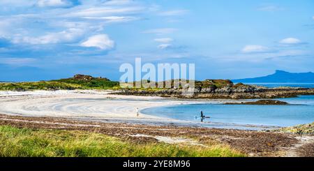 Dog walker and paddleboarder on Traigh Beach near Portnaluchaig in Lochaber, Scottish Highlands, Scotland, with Isle of Eigg in the distance Stock Photo