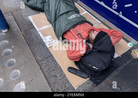 A homeless man lies in his sleeping bag on the pavement along Berwick Street Market during a working week lunchtime, Soho, London, England, UK Stock Photo