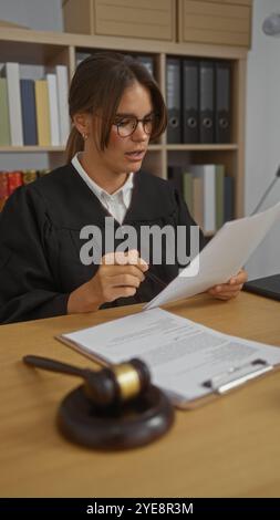 Young woman judge in courtroom reading documents with a gavel on the desk in an indoor office setting Stock Photo