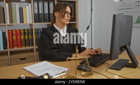 Young woman in a courtroom wearing a judge's robe working on a computer in an office setting with law books and legal scales Stock Photo