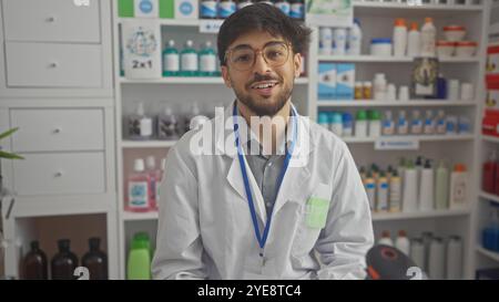 A young adult arab man with a beard wears a white lab coat and glasses, posing confidently in a pharmacy full of medicine shelves. Stock Photo