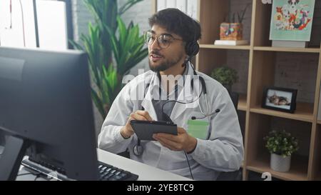 Handsome bearded man in white coat with headset and stethoscope engages with tablet in modern clinic office. Stock Photo