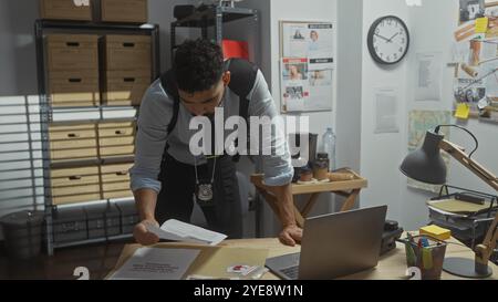 A focused hispanic man with a beard examines documents at a detective's desk cluttered with evidence and a laptop in a police station office setting. Stock Photo
