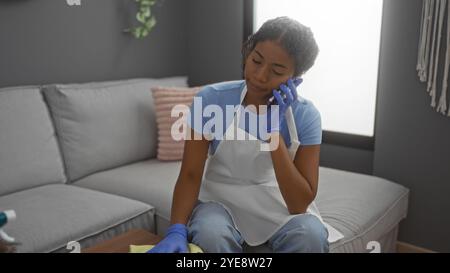 Woman wearing gloves and apron talking on the phone while cleaning a living room in her apartment. Stock Photo