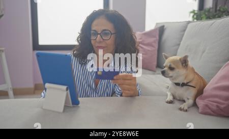 A middle-aged hispanic woman using a credit card for online shopping next to her chihuahua in a cozy living room. Stock Photo