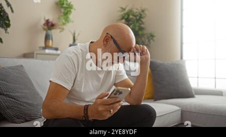 Bald man with a beard and glasses sitting in a living room holding a phone in his hand, surrounded by indoor plants and a cozy home interior Stock Photo