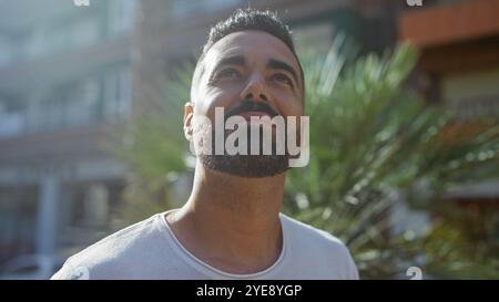 Portrait of a contemplative hispanic man looking upwards, in a sunlit urban outdoor setting with green foliage and blurred buildings. Stock Photo