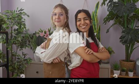 Two women, arms crossed, standing together amidst potted plants, evoke teamwork in a flower shop setting. Stock Photo