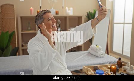 Handsome man in spa making a video call on his smartphone while wearing a white bathrobe, surrounded by wellness items in a serene beauty salon Stock Photo