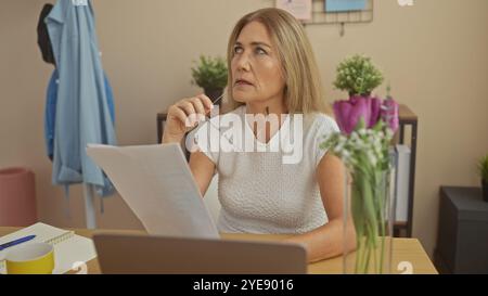 A thoughtful middle-aged woman examines documents at home, sitting with a laptop and glasses in hand, suggesting business or personal finance manageme Stock Photo