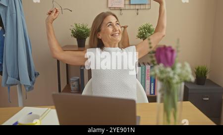 Mature woman stretching arms with a smile, relaxed ambiance in a home office with books, flowers, and laptop. Stock Photo