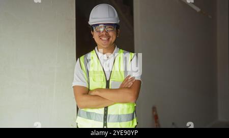 Young man wearing safety gear standing indoors with arms crossed at a construction site Stock Photo