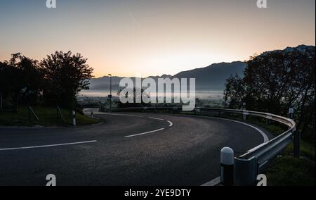 Empty curvy mountain pass at dawn. Mountain valley covered in fog in the background, sky is glowing - light coming from behind the mountains. Stock Photo