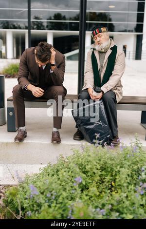 Full length vertical portrait of chatting businessman and old homeless man sitting on bench, representing growing gap between rich and poor in society Stock Photo