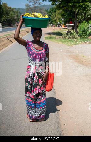 Tanzanian woman in colourful dress carries a large basket of bananas on her head; Mto wa Mbu, Tanzania Stock Photo