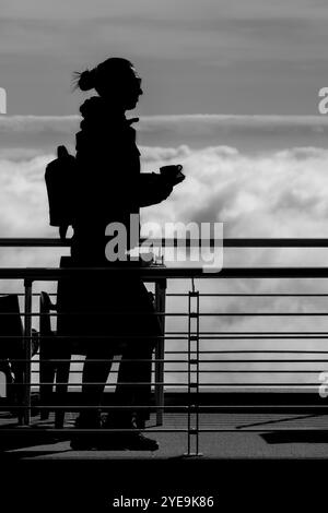 Black and white image of a silhouetted woman carrying coffee cup along walkway above the clouds at Pico do Arieiro Hike, São Roque do Faial Stock Photo