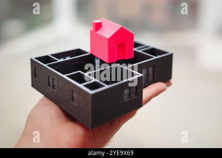 A hand displays a detailed 3D model featuring a striking red house situated on a black base, representing modern construction design and architectural Stock Photo