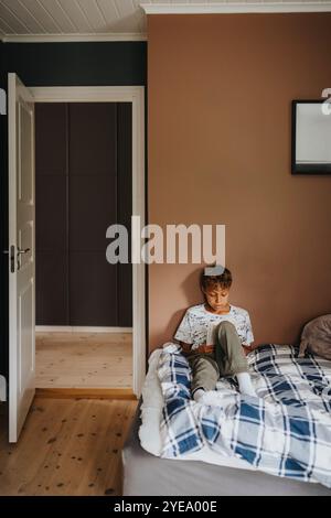 Focused multiracial boy reading book while sitting on bed in bedroom at home Stock Photo