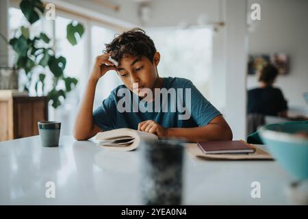 Boy leaning on elbow while reading book near table at home Stock Photo