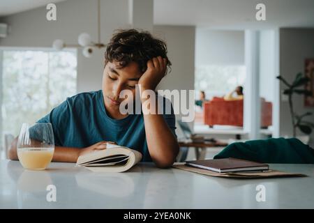 Boy leaning on elbow while reading book and sitting near table at home Stock Photo