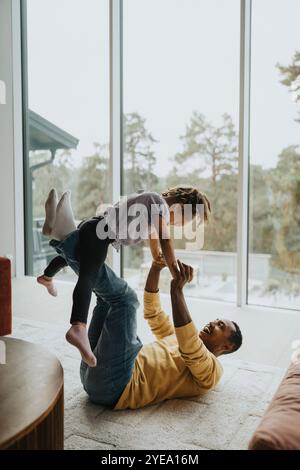 Happy man picking up daughter while lying on carpet in living room at home Stock Photo
