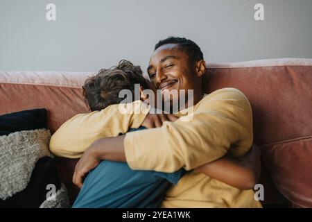 Smiling father embracing son while sitting on sofa at home Stock Photo